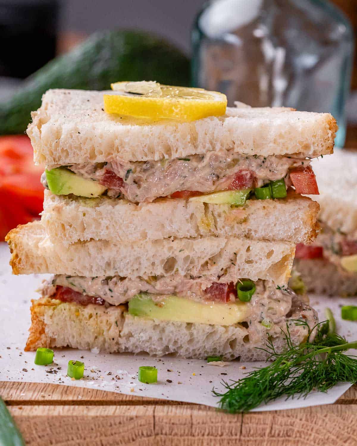 Two stacked halves of a tunacado sandwich on toasted bread, garnished with a lemon slice, fresh dill, and green onion.