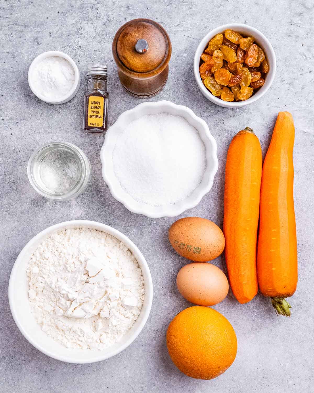 Overhead view of carrot cake ingredients laid out on a counter, including carrots, eggs, flour, sugar, raisins, orange, and vanilla.
