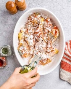 A hand drizzling olive oil over seasoned chicken drumsticks dusted with cornstarch, smoked paprika, and Italian herbs in a white baking dish, ready to roast.