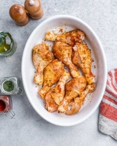 Raw chicken drumsticks evenly coated in seasoning and olive oil, arranged in a white baking dish.