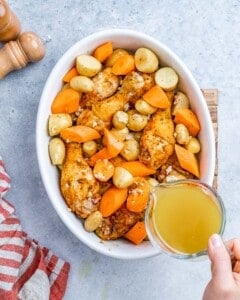 Baking dish filled with roasted drumsticks, potatoes, carrots, and onions being drizzled with chicken stock for extra flavor.