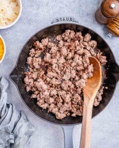 Ground beef browning in a skillet with a wooden spoon stirring.