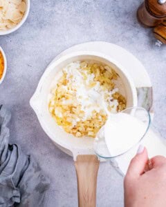 Mixing milk into shredded cheese and sautéed onions in a saucepan to make the creamy sauce.