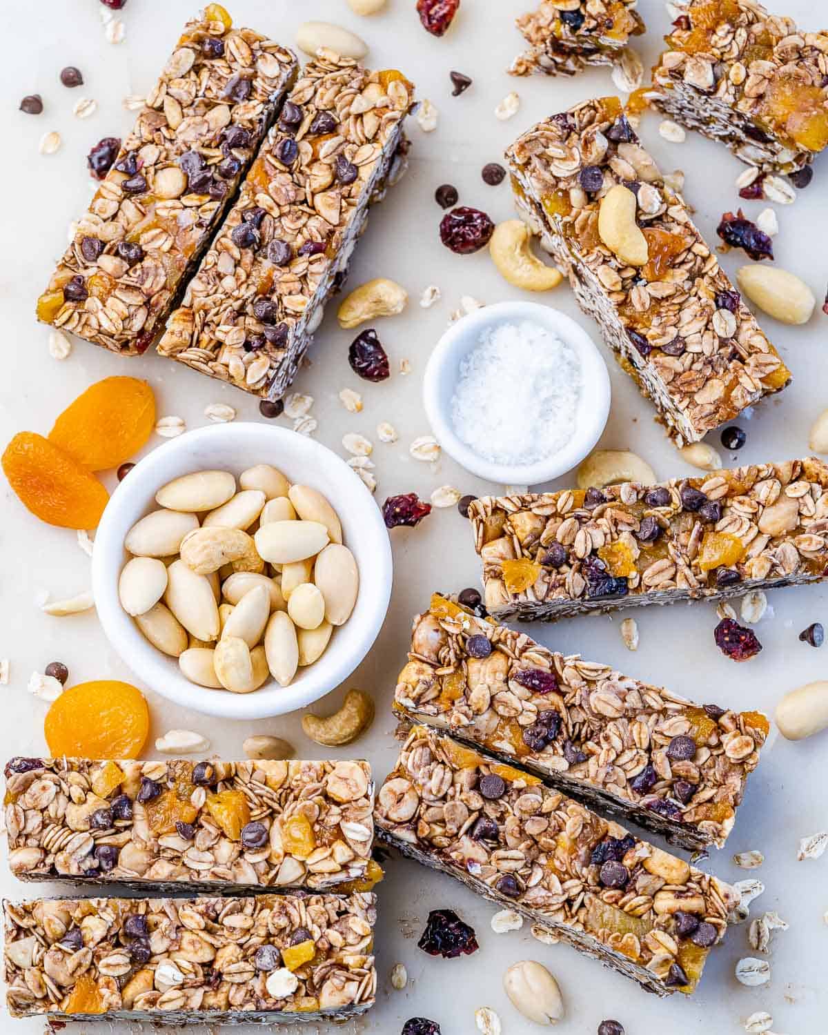 Healthy ganola bars spread out on a white surface, surrounded by bowls of dried fruit, and nuts.