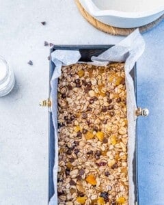 Rectangular loaf pan lined with parchment paper filled with pressed granola mixture with visible oats, nuts, dried fruit, and chocolate chips on top.