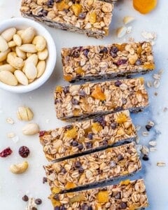 Rectangle Healthy Granola Bars in a line on a light surface next to a bowl of nuts.