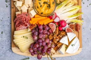 Overhead view of a colorful fruit and cheese platter featuring sliced brie, gouda, parmesan cubes, red grapes, hummus, olives, and crackers, beautifully arranged on a wooden board.