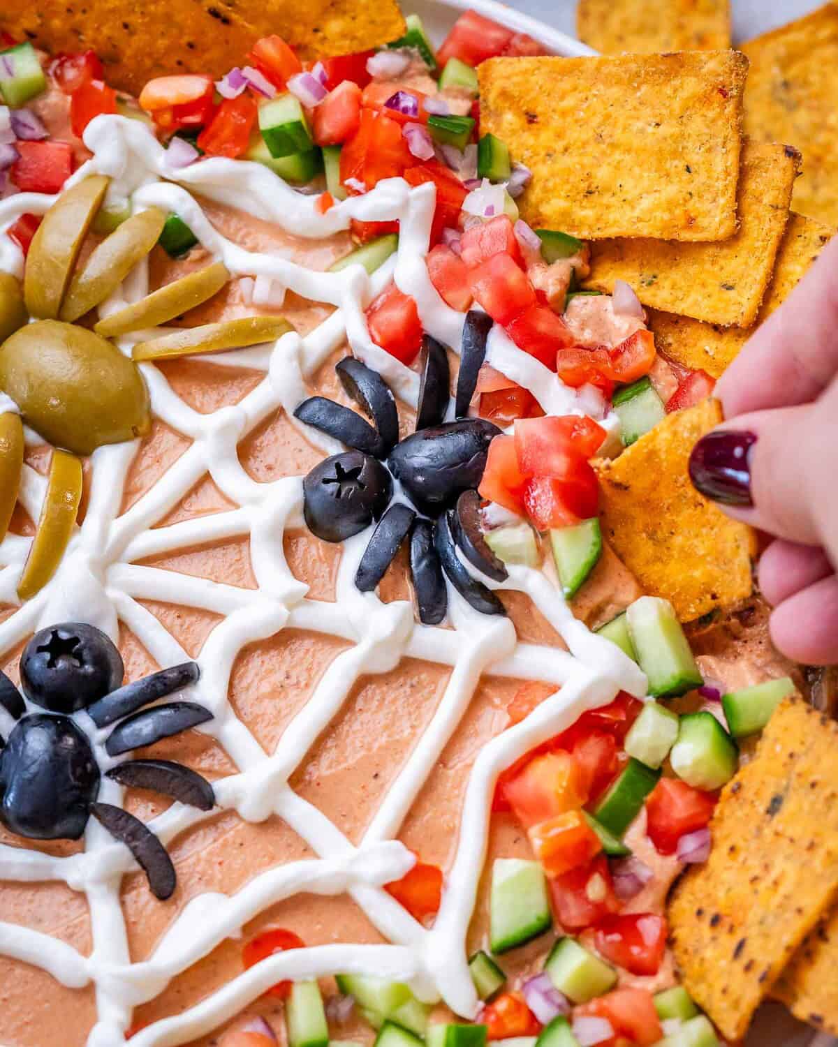 Hand grabbing a tortilla chip to scoop the dip out near a spider made out of olives on top of the sour cream spider web.
