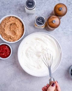 Whisk mixing the Greek yogurt, cream cheese, and seasoning together in a glass bowl.