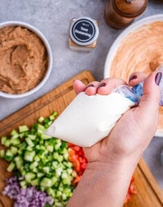 All ingredients for Halloween taco dip prepped and arranged on a cutting board and small bowls, ready to assemble, with a hand holding a piping bag full of sour cream above it.