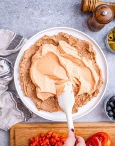 Cream cheese and Greek yogurt mixture spread over the beans in the serving dish with a white spatula.