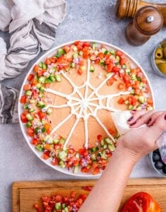 Top view of a hand piping a sour cream spider web design on top of the taco dip with diced veggies along the edges.
