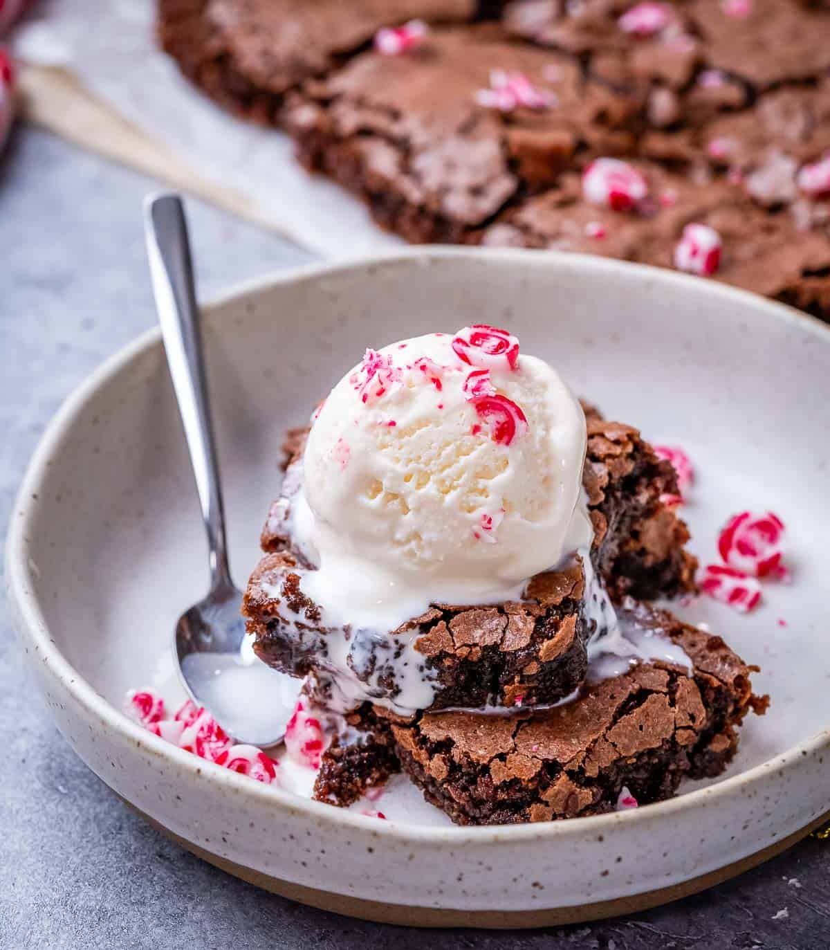 Stack of peppermint brownies in a bowl topped with vanilla ice cream and crushed peppermint candies, with a spoon beside it.