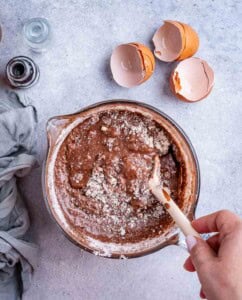 Mixing bowl with brownie batter being stirred using a wooden spoon, surrounded by cracked egg shells.