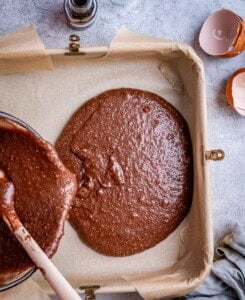 Pouring thick chocolate brownie batter into a parchment-lined square baking pan.