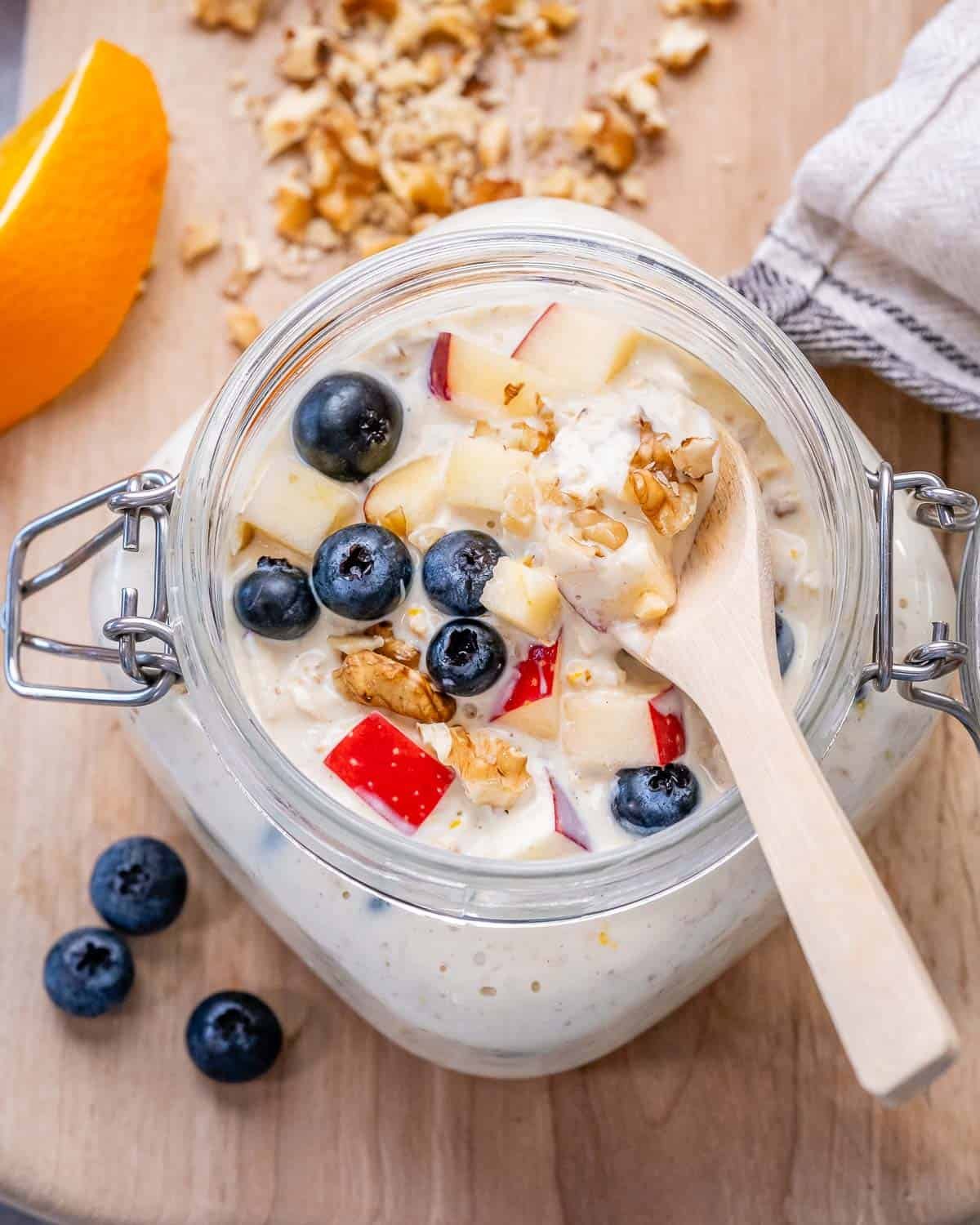 a spoon in a jar with overnight oats topped with blueberries and chopped apples.