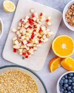 chopped apple on a cutting board, next to a bowl with oats and orange juice.