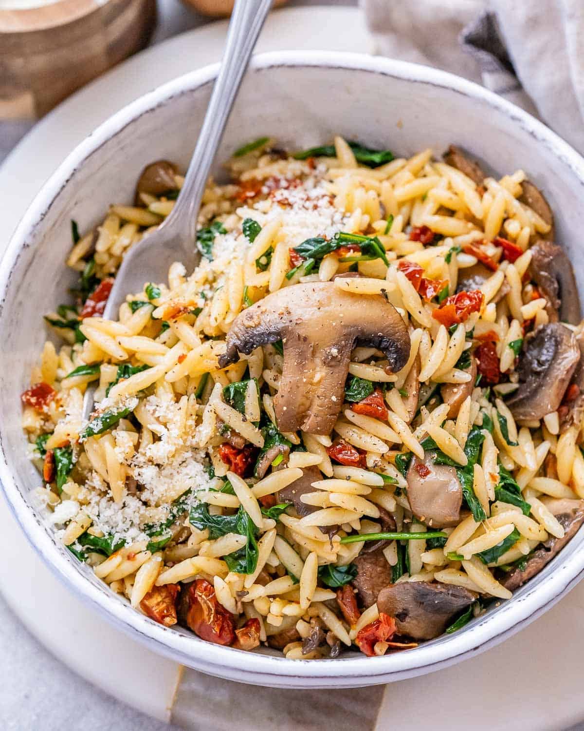 Close-up of a bowl filled with cooked orzo, mushrooms, spinach, and sun-dried tomatoes mixed together.