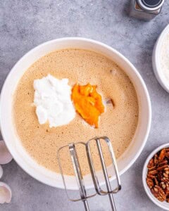Sweet potato puree and Greek yogurt being added to the egg and sugar mixture in a white bowl with an electric mixer.