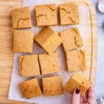 Freshly baked sweet potato cornbread cut into neat golden squares on parchment paper, with a hand holding a piece of sweet potato cornbread.