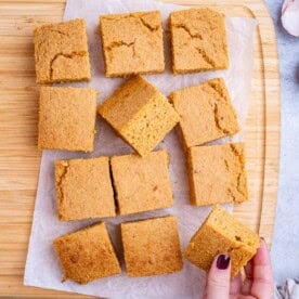 Freshly baked sweet potato cornbread cut into neat golden squares on parchment paper, with a hand holding a piece of sweet potato cornbread.