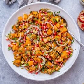 Mixed sweet potato salad in a white bowl with spoon, showing even coating of dressing and fresh herbs throughout.