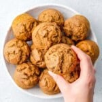 hand grabbing a single pumpkin banana muffin from a bowl filled with them.