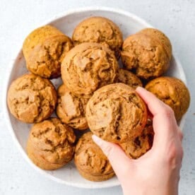 hand grabbing a single pumpkin banana muffin from a bowl filled with them.