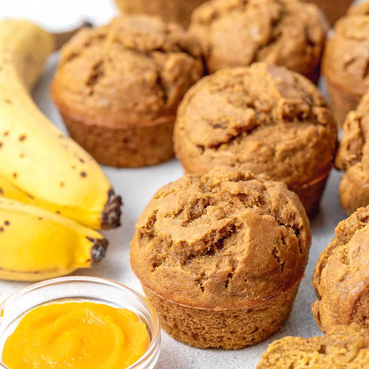 pumpkin banana muffins on a flat surface next to a small bowl of pumpkin puree and a banana.