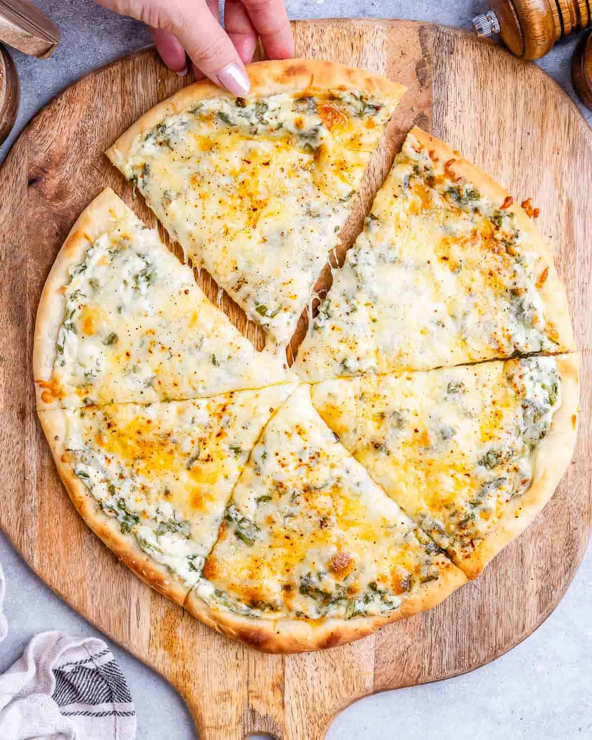 A white pizza sliced into 6 slices on a wooden board with a hand pulling one slice away.