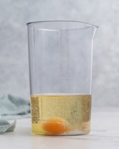 All ingredients combined in a tall glass jar before blending: whole egg at the bottom, Dijon mustard, lemon juice, vinegar, salt, and avocado oil poured on top.