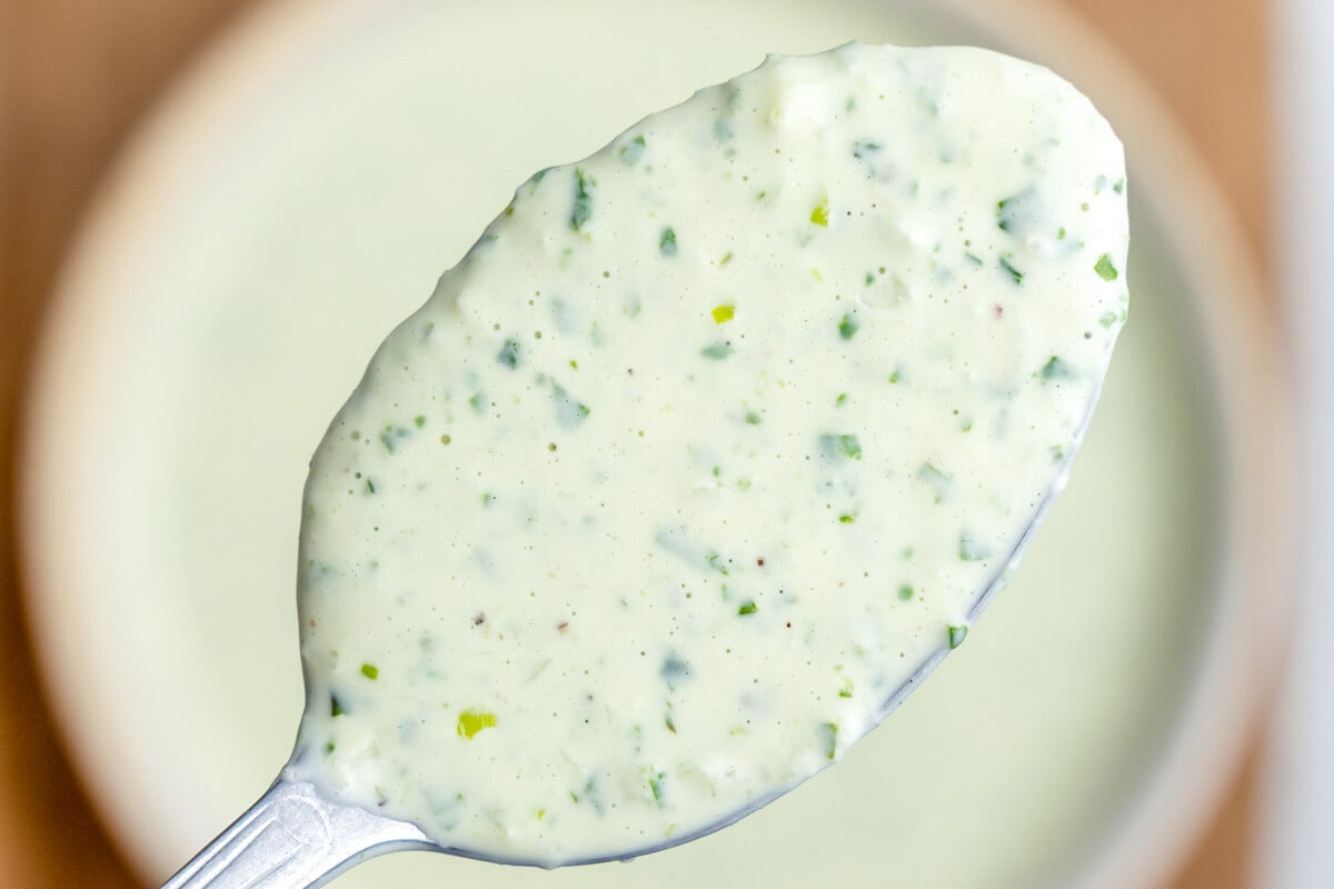 A closeup of a spoonful of green goddess dressing hovering over a bowl.