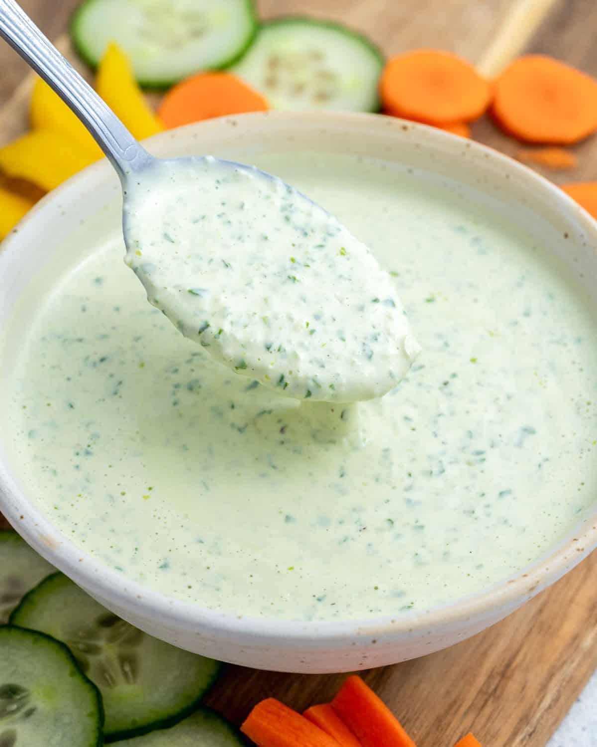 A white bowl of green goddess dressing being scooped with a spoon, surrounded by fresh vegetables. 