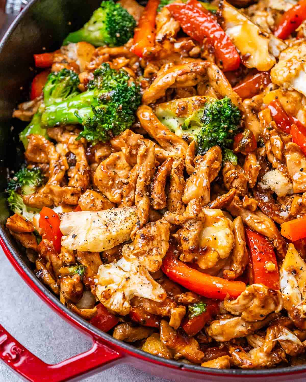 Another close-up of the finished stir-fry in the pan, highlighting the glossy sauce, browned chicken strips, red bell peppers, broccoli florets, and cauliflower pieces.