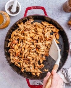 Action shot in a large red-handled pan: stir-frying marinated chicken pieces with sliced red bell peppers, broccoli florets, and cauliflower, with a white spatula tossing the ingredients.