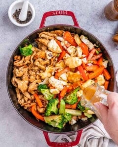 Stir-fry in progress in a large pan on a gray surface: tender chicken pieces, red bell pepper strips, broccoli, and cauliflower florets being cooked together with visible glossy sauce beginning to coat everything.