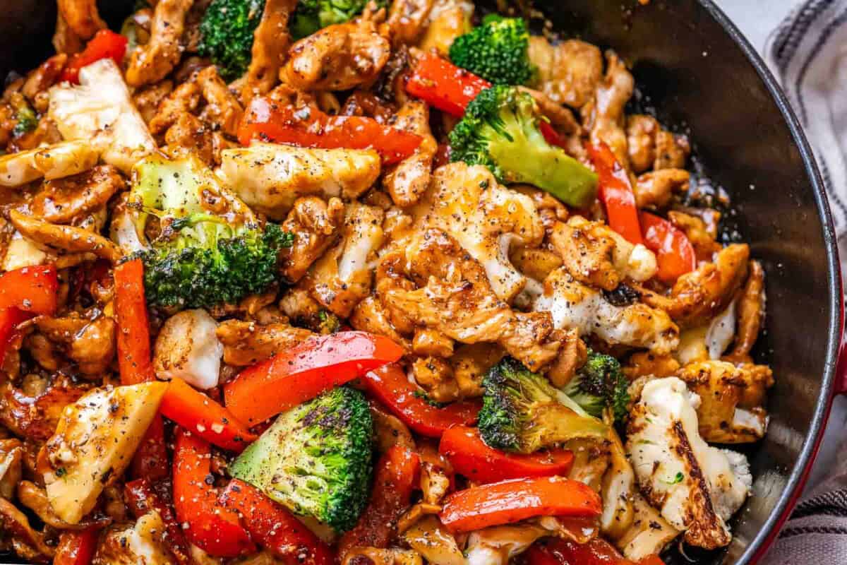 Close-up of plated black pepper chicken stir-fry with tender chicken pieces, red bell pepper strips, broccoli, and cauliflower all coated in thick, shiny black pepper sauce.