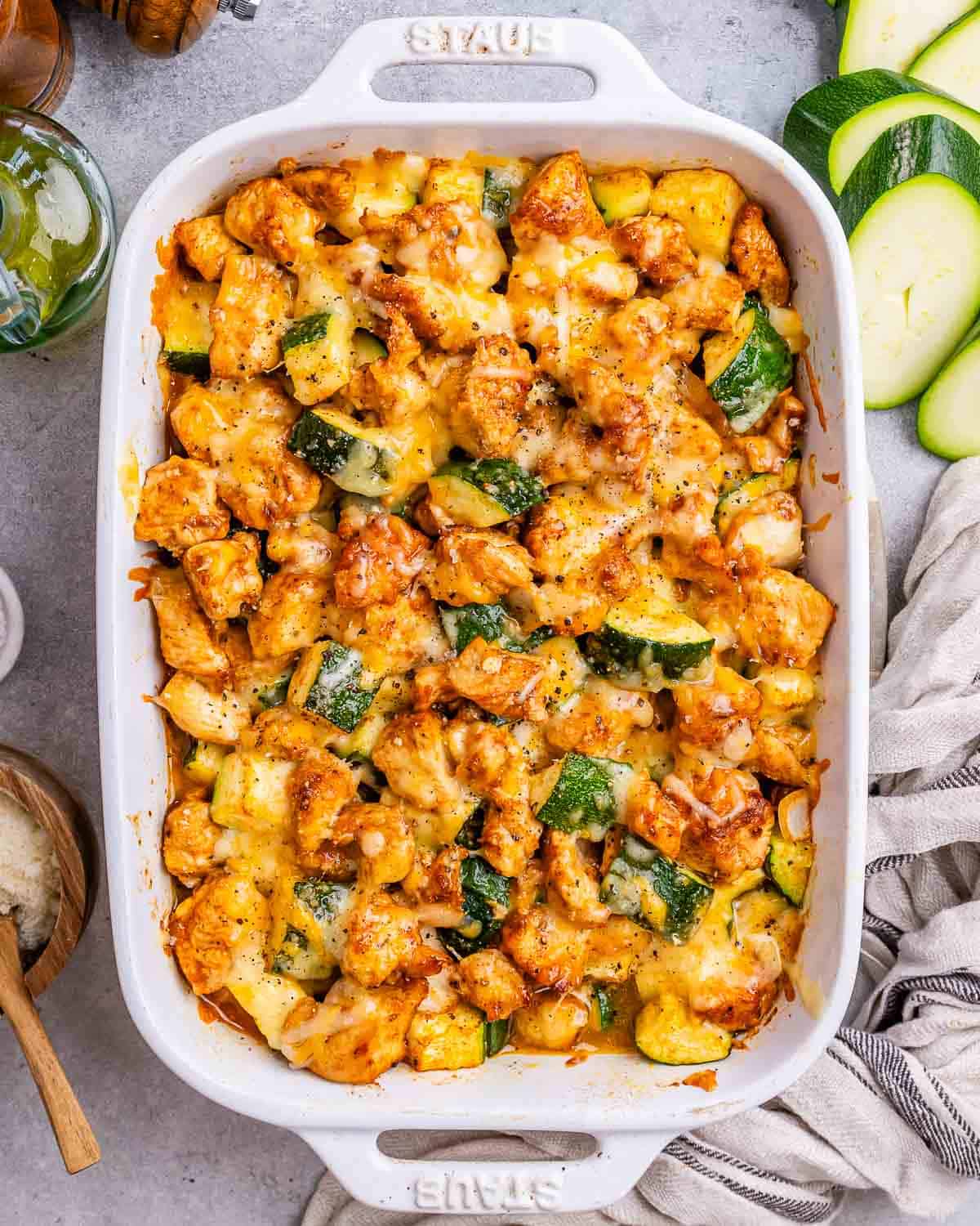 a white baking dish with baked chicken and zucchini bites topped with cheese.