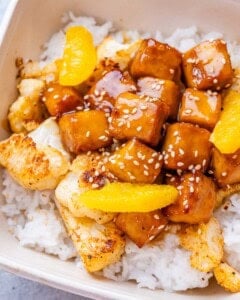 A close-up image of crispy orange tofu and cauliflower with rice in a white bowl.