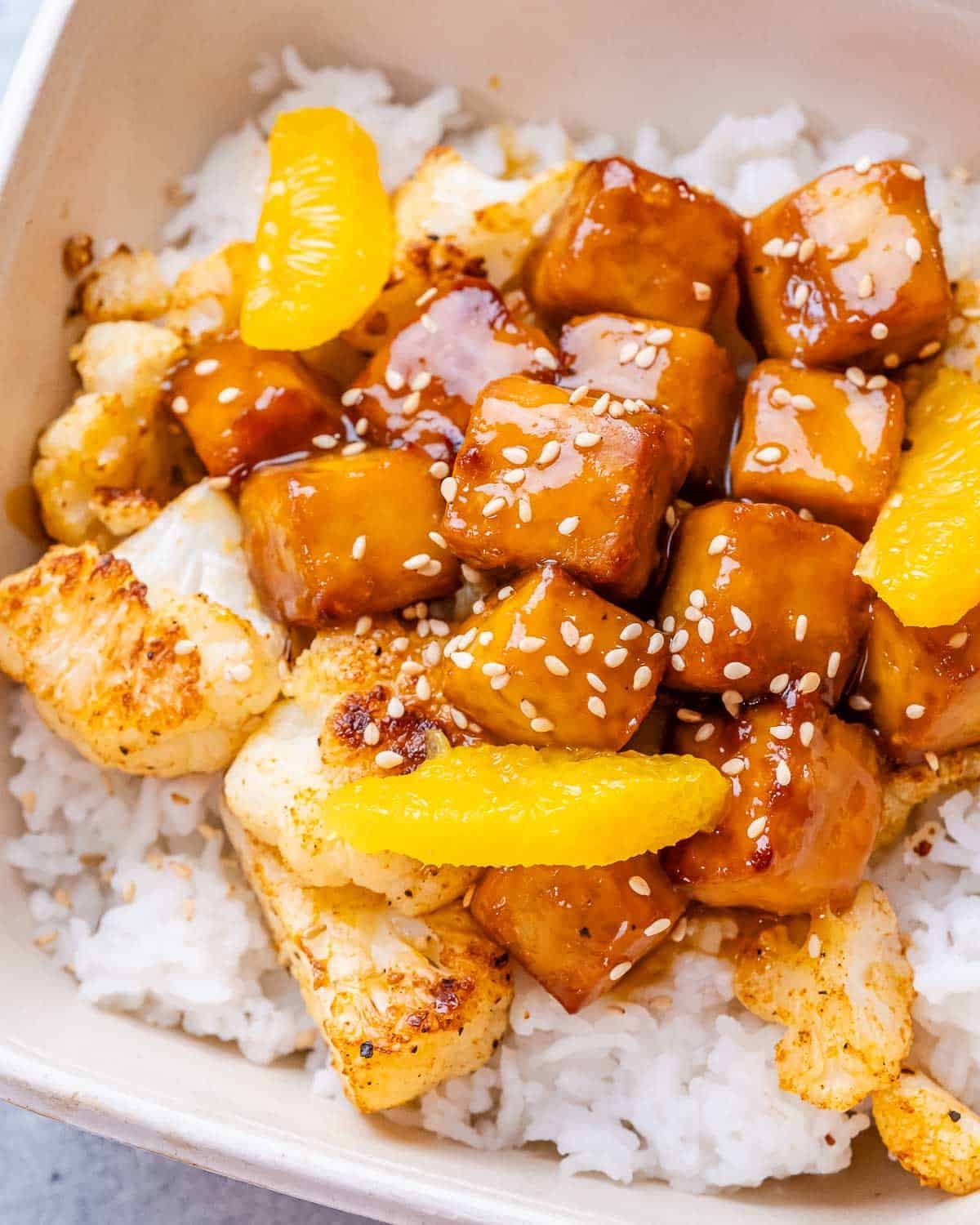 A close-up image of crispy orange tofu and cauliflower with rice in a white bowl.