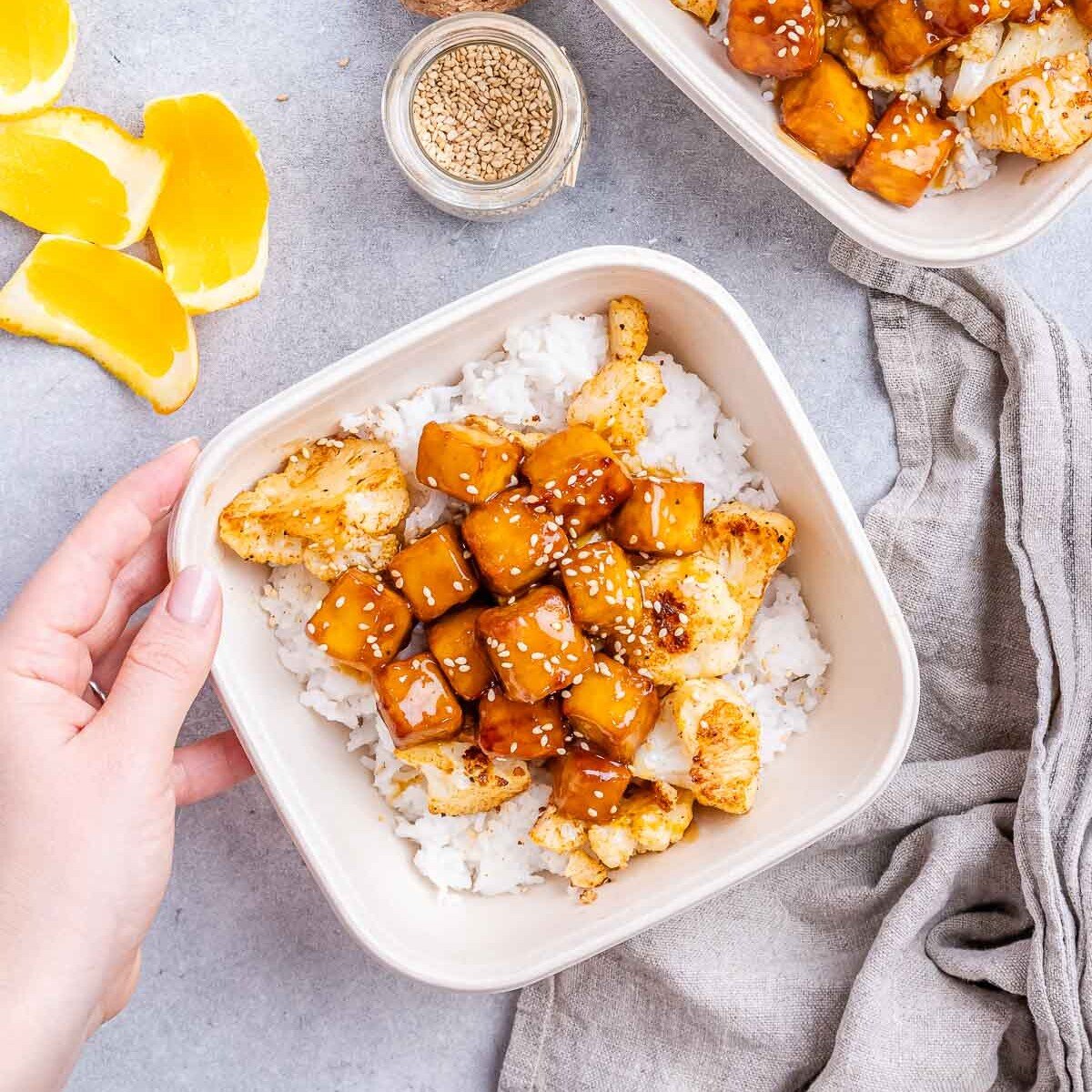 A hand holding a meal prep bowl filled with rice and crispy orange tofu.