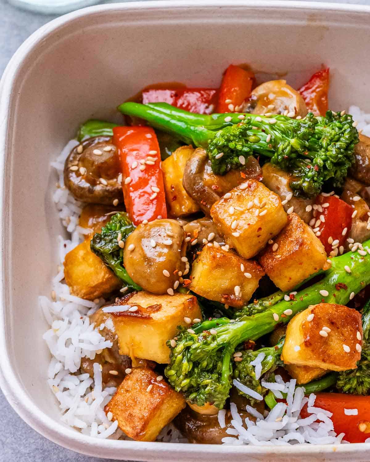 Top view close up of a white square bowl with rice atop with tofy stir fry.