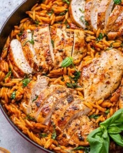 Top view of the completed dish in a red Dutch oven with juicy sliced Tuscan chicken breasts resting over vibrant orzo pasta mixed with spinach, sun-dried tomatoes, and fresh basil.