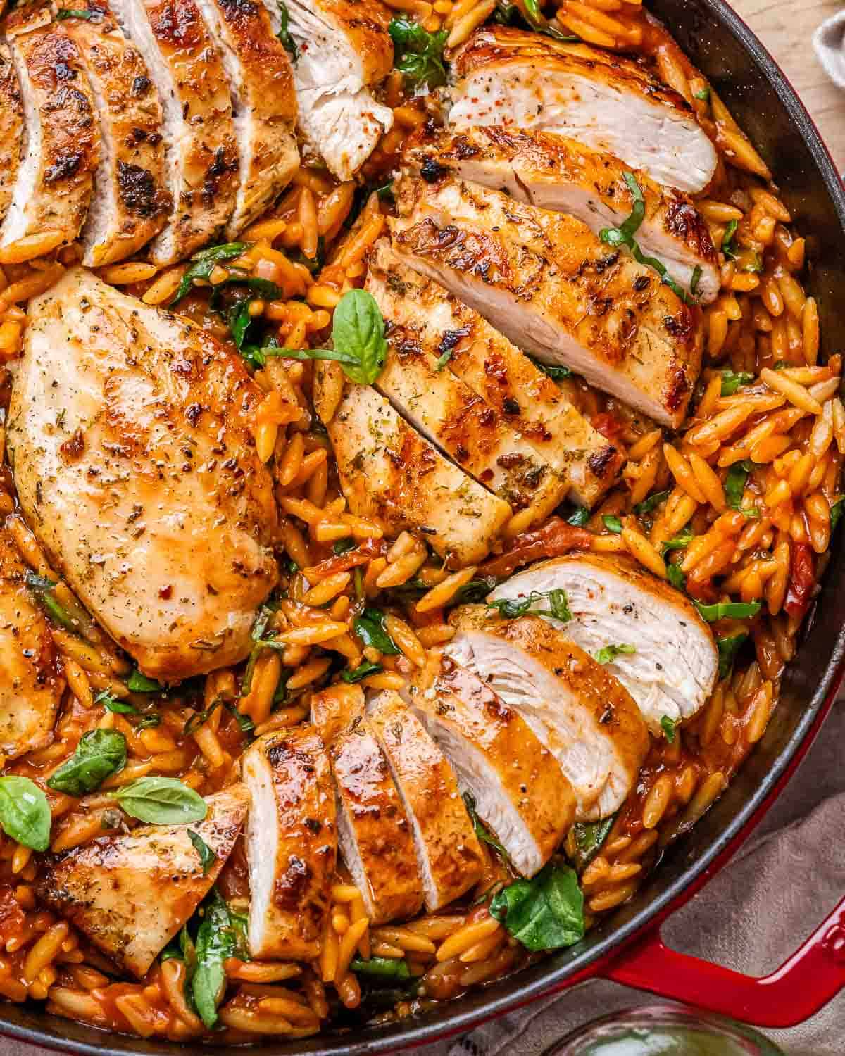 Close-up of the completed dish in a red Dutch oven with juicy sliced Tuscan chicken breasts resting over vibrant orzo pasta mixed with spinach, sun-dried tomatoes, and fresh basil.