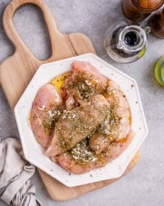 Seasoned raw chicken breasts coated in Italian seasoning and garlic powder, resting on a white octagonal plate with a wooden board of fresh basil and spinach in the background.