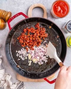 Close-up of saut&eacute;ing chopped sun-dried tomatoes, diced shallots, minced garlic, and red pepper flakes in oil inside a black skillet.