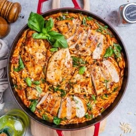 A red skillet with tomato based orzo topped with seared sliced chicken.