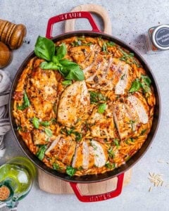 Top view of creamy Tuscan Chicken Orzo in a large red Dutch oven, showing sliced chicken on top of orzo mixed with wilted spinach, sun-dried tomatoes, and fresh basil garnish.