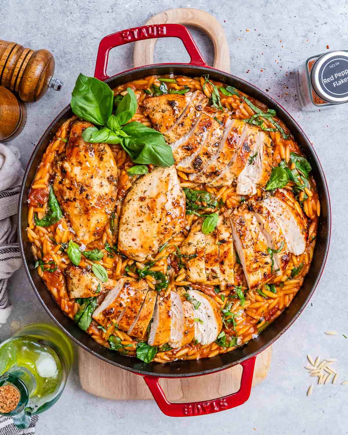 Top view of creamy Tuscan Chicken Orzo in a large red Dutch oven, showing sliced chicken on top of orzo mixed with wilted spinach, sun-dried tomatoes, and fresh basil garnish.