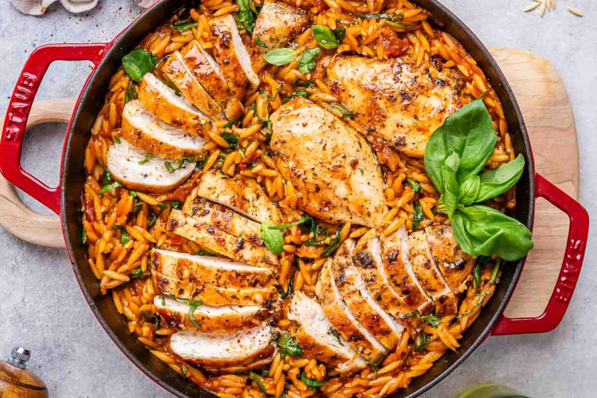 Close up of sliced cooked chicken breasts arranged over a pan of creamy Tuscan orzo pasta tossed with sun-dried tomatoes, spinach, and herbs.